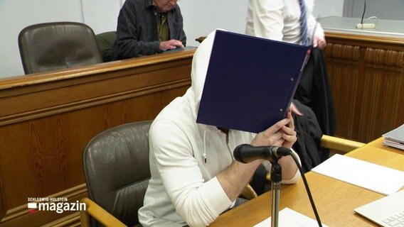 Eine Person in einem Gerichtssaal hält sich einen Aktenordner vors Gesicht. © Screenshot 