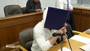 Eine Person in einem Gerichtssaal hält sich einen Aktenordner vors Gesicht. © Screenshot 