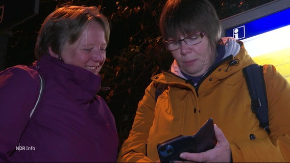 Zwei Personen an einem Bahnsteig schauen auf ihr Smartphone. © Screenshot 