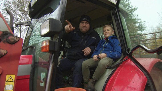 Ein Mann und ein Junge sitzen auf einem roten Trecker, der Mann zeigt mit ausgestrecktem Arm auf etwas außerhalb des Bildes. © Screenshot 