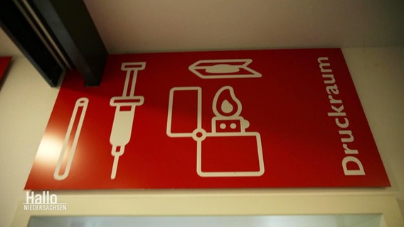Ein rotes Schild mit dem Schriftzug "Druckraum" im "Stellwerk Hannover", einer Schutz- und Sozialeinrichtung für Drogenabhängige. Abgebildet sind auch noch eine Spritze und ein Feuerzeug. © Screenshot 