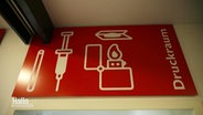 Ein rotes Schild mit dem Schriftzug "Druckraum" im "Stellwerk Hannover", einer Schutz- und Sozialeinrichtung für Drogenabhängige. Abgebildet sind auch noch eine Spritze und ein Feuerzeug. © Screenshot 