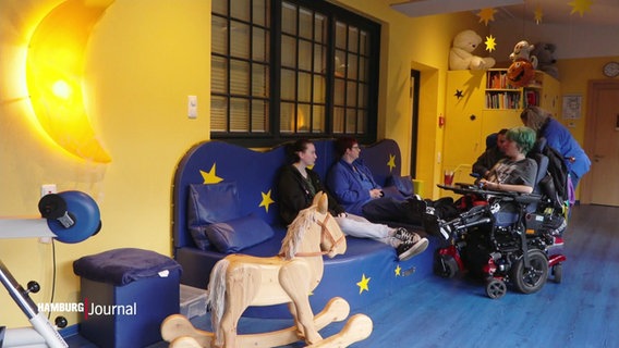 Mehrere Menschen im Kinder-Hospiz Sternenbrücke: Zwei Personen sitzen auf einer großen, blauen Couch, zwei Personen sitzen in Rollstühlen. An der Wand hängen gelbe Sterne, im Vordergrund steht ein Schaukelpferd. © Screenshot 