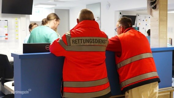 Zwei Mitarbeiter des Rettungsdienstes in oranger Arbeitskleidung von hinten zu sehen. Sie lehen an einem Empfangstresen in einer Klinik. © Screenshot 
