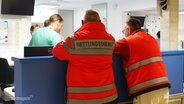 Zwei Mitarbeiter des Rettungsdienstes in oranger Arbeitskleidung von hinten zu sehen. Sie lehen an einem Empfangstresen in einer Klinik. © Screenshot 