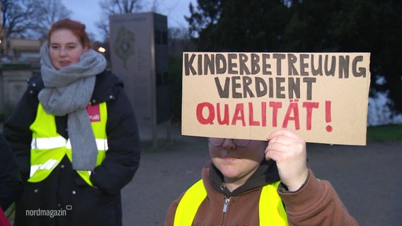 Auf einer Mahnwache  hält eine Erzieherin ein Schild auf dem "Kinderbetreuung verdient Qualität!" steht. © Screenshot 