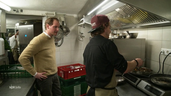Zwei Männer in einer Gastro-Küche: Einer steht am Herd und kocht, der andere sieht im dabei zu und spricht mit ihm. © Screenshot 
