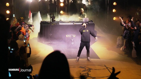 Bei einem Dance-Battle spricht ein Kapuzenpulli-tragender Mann vor einem DJ-Pult in ein Mikrophon, während die Zuschauenenden wild jubeln. © Screenshot 