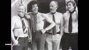 Vier Männer posieren mit freiem Oberkörper und um den Hals gebundenen Krawatten auf einem älteren Schwarz--Weiß-Foto. © Screenshot 