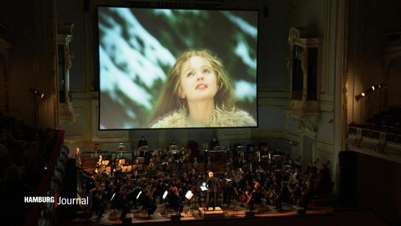 Vor einer riesigen Leinwand auf die der Film "Drei Haselnüsse für Aschenbrödel" projiziert wird spielt ein Orchester in der Laeiszhalle. © Screenshot 