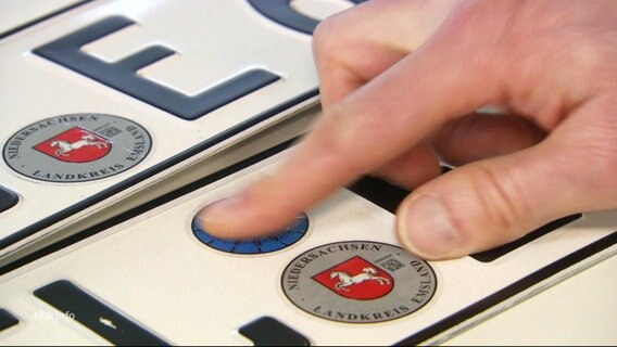Nahaufnahme: Beamter klebt TÜV-Plakette auf ein Auto-Kennzeichen. © Screenshot 