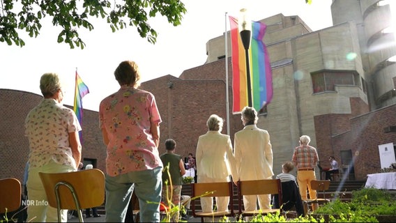 Lesbische Paare stehen vor einer Kirche und blicken zu einer gehissten Regenbogenflagge. © Screenshot 