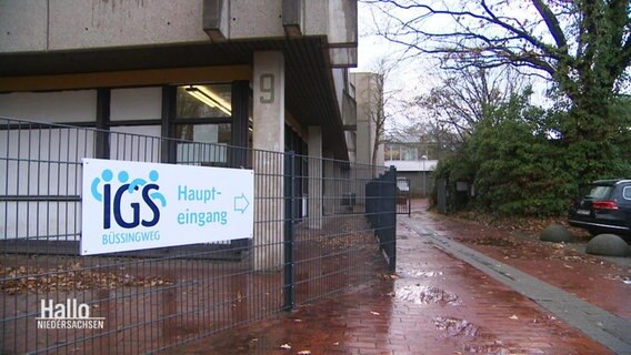 Blick auf ein Schulgebäude der IGS Büssingweg. Ein Zaum führt um das Gebäude herum, auf dem ein Schuld montiert ist: IGS Büssingweg, mit einem Pfeil Richtung Haupteingang. © Screenshot 
