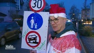Ein Mann steht mit einem selbst gebastelten Verkehrsschild in der Hand an einer Straße. Er trägt eine Weihnachtsmann-Mütze und einen rot-weiß-gestreiften Signalanzug. © Screenshot 