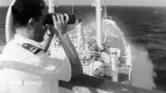 In einer alten Archivaufnahme blickt ein Mann auf einem Schiff durch ein Fernglas. © Screenshot 