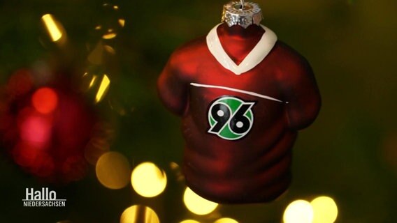 Ein Weihnachtsanhänger in Form eines Fußball-Trikots mit dem Logo von Hannover 96 hängt an einem Tannenzweig. © Screenshot 