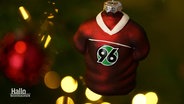 Ein Weihnachtsanhänger in Form eines Fußball-Trikots mit dem Logo von Hannover 96 hängt an einem Tannenzweig. © Screenshot 