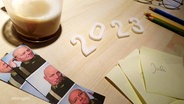 Nahaufnahme eines Schreibtischs auf dem die Jahreszahl "2023", bestehend aus Pappe, neben einem Notizblock, Stiften und Drucke aus einem Fotoautomaten drapiert herumliegen. © Screenshot 