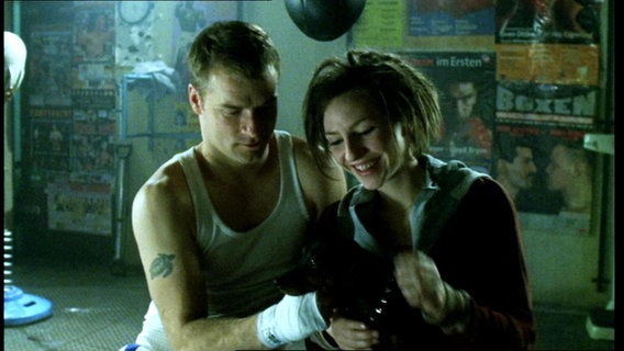 Ein junger Mann im Unterhemd und mit Tattoo auf dem Oberarm und eine junge Frau mit Dreadlocks und Kapuzenpulli streicheln einen kleinen, schwarzen Hund. Im Hintergrund Poster von Boxkämpfen. © Screenshot 