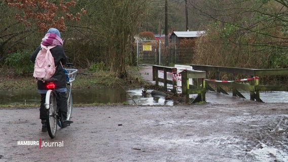 Eine Radfahrerin steht vor einer durch das Hochwasser gefährdeten, abgesperrten Brücke. © Screenshot 