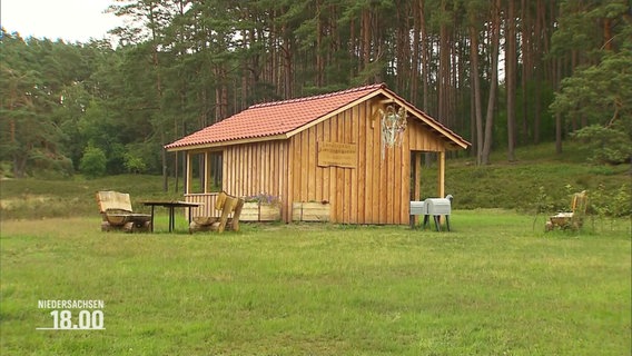 EIne aus Holz gebaute Schutzhütte auf einer grünen Wiese am Waldrand. Davor ein paar Bänke und Tische. © Screenshot 