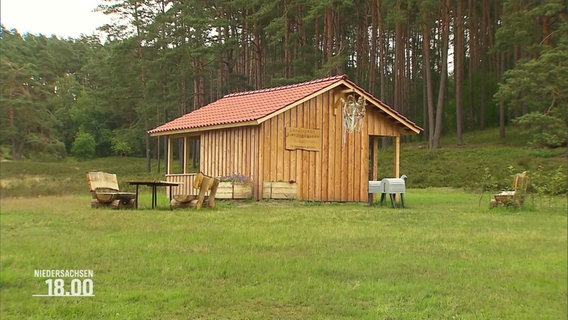 EIne aus Holz gebaute Schutzhütte auf einer grünen Wiese am Waldrand. Davor ein paar Bänke und Tische. © Screenshot 