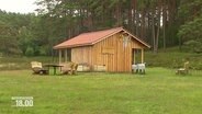 EIne aus Holz gebaute Schutzhütte auf einer grünen Wiese am Waldrand. Davor ein paar Bänke und Tische. © Screenshot 