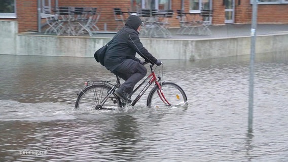 Eine Person fährt bei Hochwasser Fahrrad, wobei dasWasser bis zur Radnabe reicht. © Screenshot 