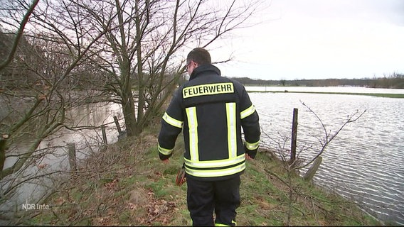 Mitglieder der Freiwilligen Feuerwehr gehen zur Kontrolle auf einem Deich entlang. Das Wasser steht links und rechts des Deiches sehr hoch. © Screenshot 