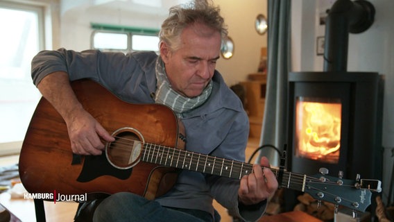 Werner Pfeifer spielt Gitarre auf seinem Hausboot. Im Hintergrund ein Feuer in einem Ofen. © Screenshot 