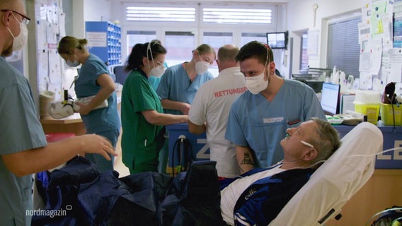 Szene in einer Klinik: Im Vordergrund ein Patient auf einer Liege, neben ihm ein Krankenpfleger, im Hintergrund Sanitäter und medizinisches Personal. © Screenshot 