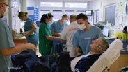 Szene in einer Klinik: Im Vordergrund ein Patient auf einer Liege, neben ihm ein Krankenpfleger, im Hintergrund Sanitäter und medizinisches Personal. © Screenshot 