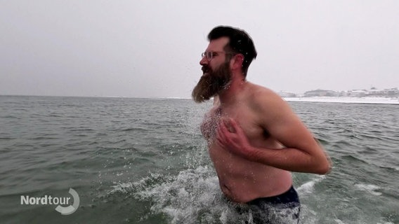 Ein Mann mit Bart und Brille steht bis zum Bauch in der eiskalten Ostsee (4 Grad). Er macht seinen Oberkörper mit den Armen nass. © Screenshot 