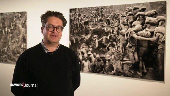 Ein Mann mit Brille steht vor Schwarz-Weiß-Fotografien in einer Ausstellung © Screenshot 