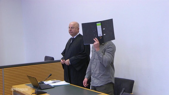 Ein Angeklagter steht in einem Gerichtssaal neben seinem Verteidiger. © Screenshot 
