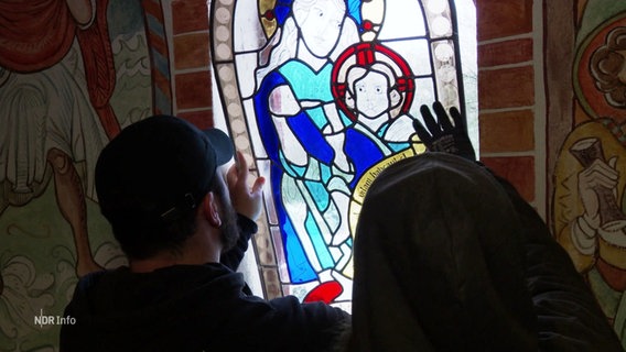 Zwei Menschen setzen ein neues buntes Fenster in eine Klostermauer ein. © Screenshot 