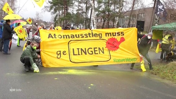 Teilnehmende einer Protestaktion halten ein gelbes Banner mit der Aufschrift: "Atomausstieg muss geLingen". Eine Person streut ein gelbes Pulver auf die Straße. © Screenshot 