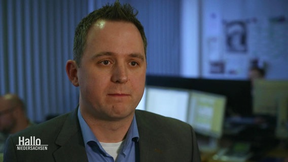 Ein Mann mit dunklen Haaren steht in einem Büro. Im Hintergrund sind Monitore zu sehen. © Screenshot 