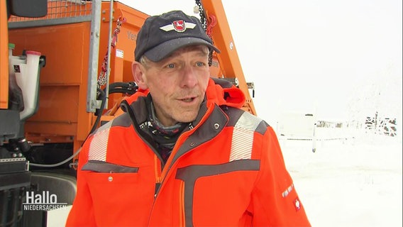 Ein Mann vom Schneeräumdienst im orangefarbener Jacke steht vor seinem Räumfahrzeug. © Screenshot 