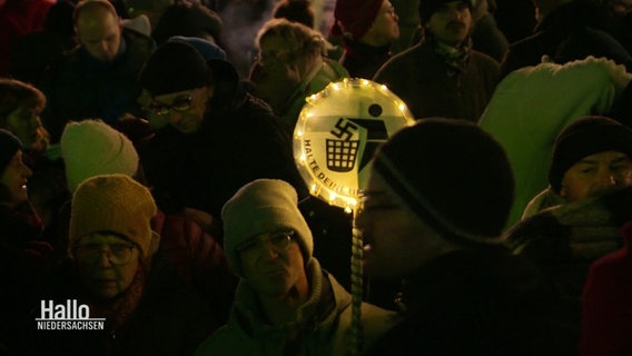 Szene auf einer Demo: Eine Person mit einem runden Schild, umkränzt von einer Lichterkette. Auf dem Schild sieht man ein Piktogramm, in dem ein Männchen ein Hakenkreuz in einen Abfalleimer wirft. Darunter die Aufschrift: "Halte deine Umwelt sauber." © Screenshot 