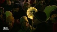 Szene auf einer Demo: Eine Person mit einem runden Schild, umkränzt von einer Lichterkette. Auf dem Schild sieht man ein Piktogramm, in dem ein Männchen ein Hakenkreuz in einen Abfalleimer wirft. Darunter die Aufschrift: "Halte deine Umwelt sauber." © Screenshot 