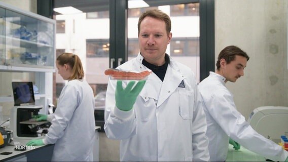 Ein Mann im weißen Kittel und mit grünen Latexhandschuhen in einem Labor, er hält eine Petrischale mit einem Fischfilet in der ausgestreckten Hand. Im HIntergrund arbeiten zwei weitere Personen. © Screenshot 