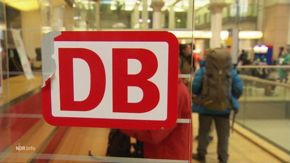 Ein Aufkleber mit dem Logo der Deutschen Bahn auf einer Glasscheibe, die linke obere Ecke fehlt. Im Hintergrund unscharf mehrere Reisende mit Gepäck. © Screenshot 