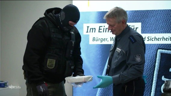 Zwei Zollbeamte übergeben sich ein Päckchen mit Rauschgift. Beide sind in Unifrom, die eine Person in schusssicherer Weste und Vermummung. © Screenshot 