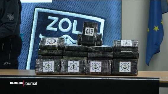 EIn Stapel abgepackter Drogen in schwarzer Folie, darauf das Symbol eines Ankers und eines Steuerrades, im Hintergrund das Wappen vom Zoll. © Screenshot 