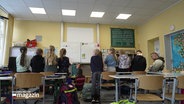 Szene in einem Klassenraum einer Grundschulklasse: Eine Lehrerin spricht mit den Schülerinnen und Schülern, die Grundschulkinder stehen nebeneinander dicht an der Tafel. © Screenshot 