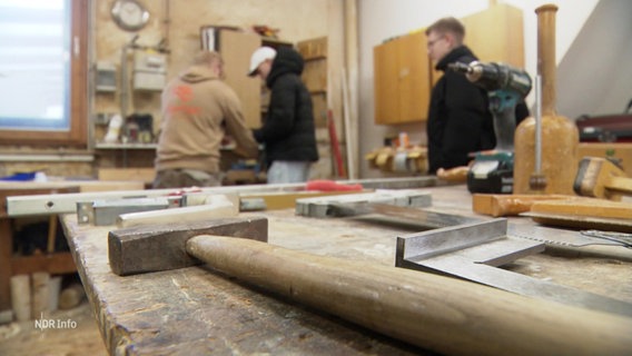 In einer Tischlerwerkstatt: Nahaufnahme eines Hammers auf einer Werkbank, im Hintergrund unscharf Jugendliche und ein Mitarbeiter der Werkstatt. © Screenshot 