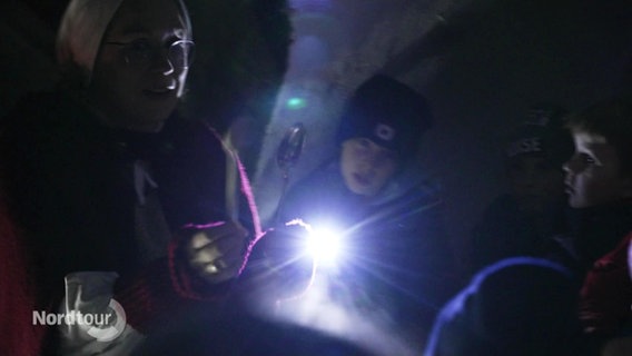 Ein Junge leuchtet mit einer Taschenlampe einen kleinen silbernen Löffel an, den eine Frau in der Hand hält. Darum herum weitere Kinder, es ist dunkel. © Screenshot 