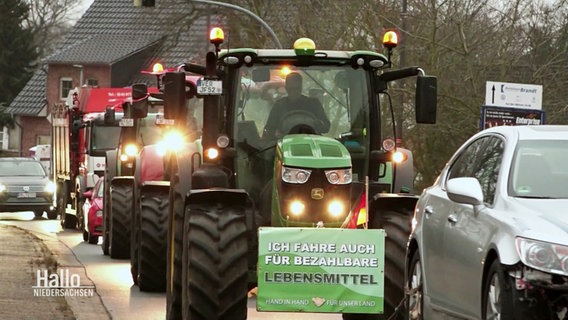 Eine Reihe von Traktoren fährt auf einer Straße, am vordersten Trecker ein Schild mit der Aufschrift: "Ich fahre auch für bezahlbare Lebensmittel". © Screenshot 