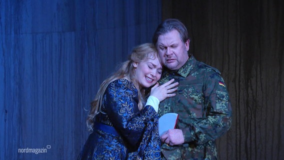 Auf einer Theaterbühne legt eine Schauspielerin ihren Kopf auf die Brust eines Mannes, der eine Bundeswehr-Uniform trägt. © Screenshot 